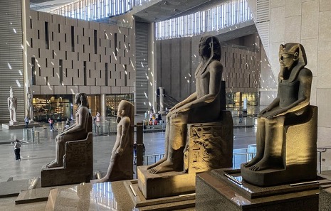 A view of sunlight pouring into the Grand Egyptian Museum's main hall, illuminating a row of colossal statues of pharaohs seated in regal poses at the Great Egyptian Museum